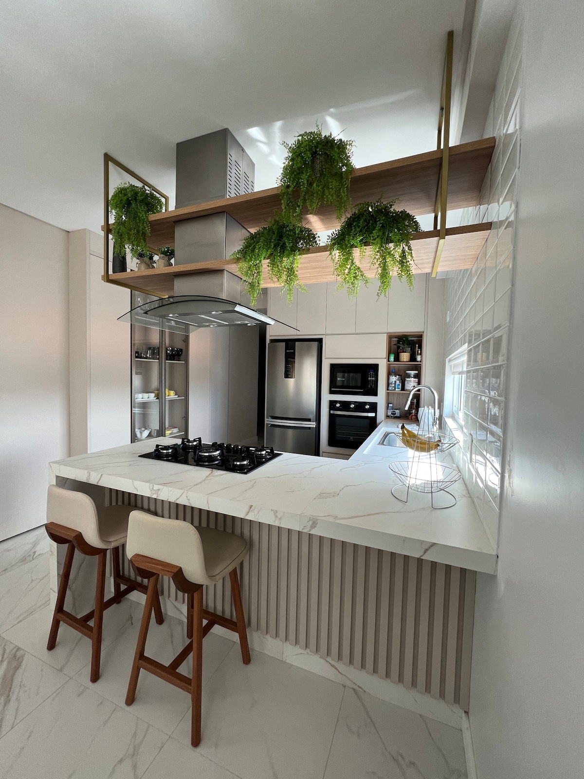 A modern kitchen showcases a sleek marble island with two barstools. Above, a wooden shelf displays lush green plants. Stainless steel appliances, including a refrigerator and an oven, are integrated into the cabinetry, enhancing the kitchen's functional design.