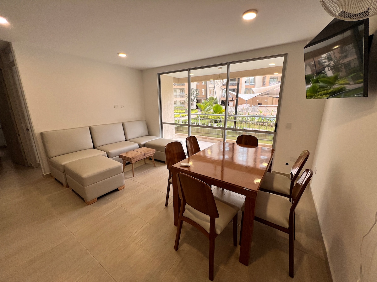 A modern living area features a light-colored sectional sofa and a wooden dining table surrounded by several chairs. Large glass doors open to a patio area adorned with greenery, while a flat-screen TV is mounted on the wall.