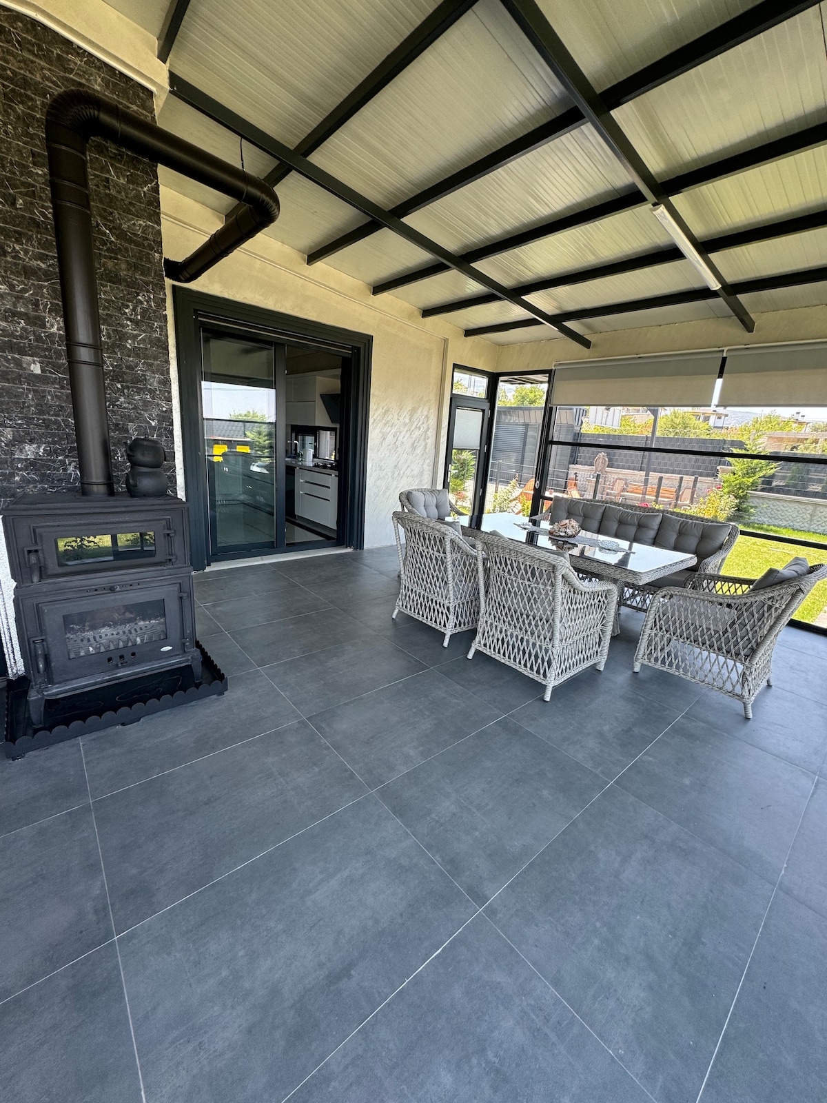 A covered outdoor dining area features a glass-top table surrounded by four comfortable wicker chairs. A wood-burning stove adds warmth, while large windows provide views of the garden. The gray floor tiles enhance the modern aesthetic of the space.