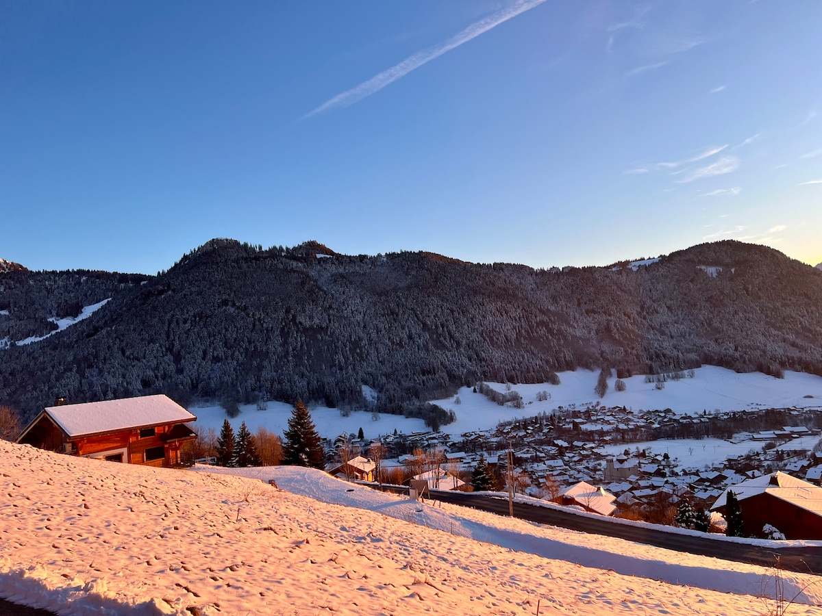 A serene winter landscape showcases rolling snowy hills and distant mountains. Sunlight casts a warm glow on the white terrain, while charming wooden chalets dot the foreground. A peaceful village can be seen nestled in the valley below, enhancing the tranquil scene.