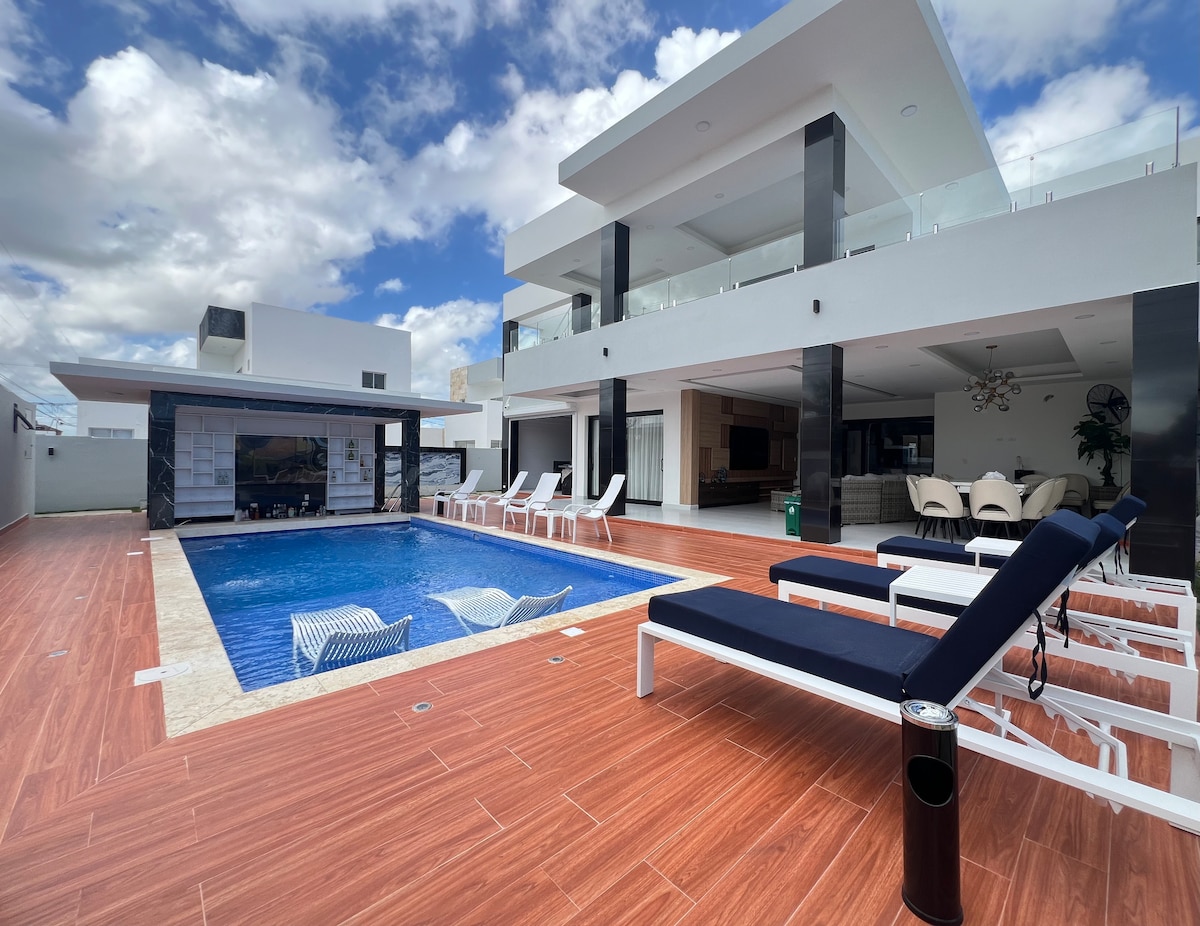 An outdoor space is featured with a large swimming pool surrounded by sun loungers and a relaxed seating area. The modern two-story building presents large windows and an open layout, allowing natural light to fill the area. Lush skies create a serene backdrop.