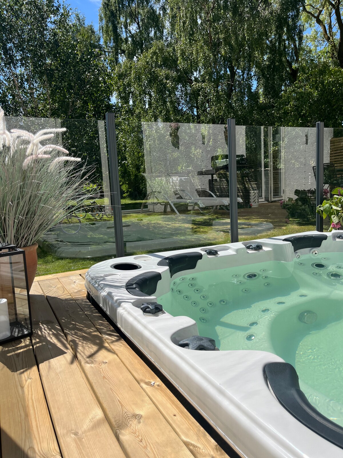 A private hot tub is set on a wooden deck, surrounded by glass panels for safety. Lush greenery and a garden area can be seen in the background, complete with comfortable seating and sunlight filtering through the trees.