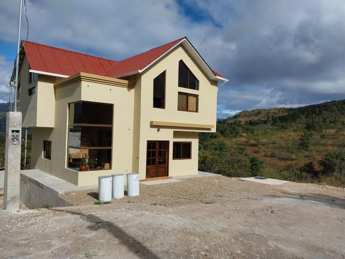 A two-story yellow house features large windows that provide ample natural light and scenic views of the surrounding hills. A red roof complements the building, while a gravel pathway leads to the entrance, offering a welcoming approach.