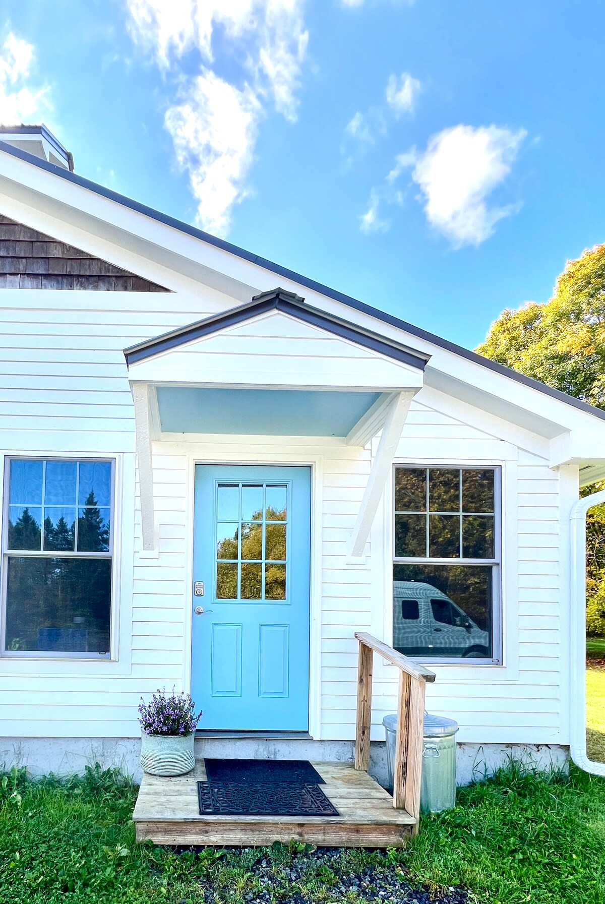 The exterior features a bright blue door framed by white siding and a small overhang. Two large windows on either side allow natural light to illuminate the entryway, while a wooden step leads onto a grassy area with a small potted plant near the entrance.