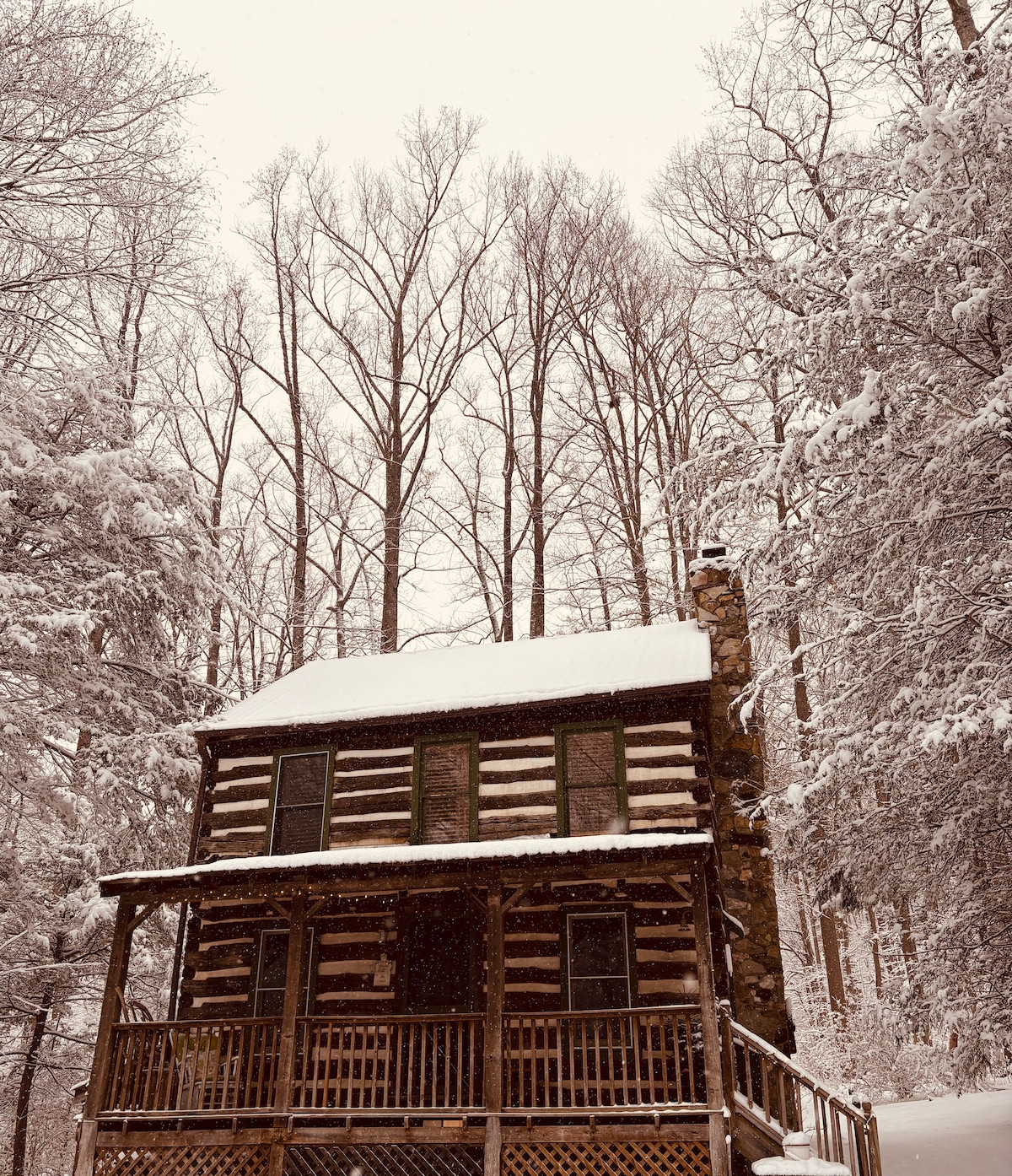 A traditional log cabin is nestled among tall, snow-covered trees. The structure features a covered front porch and a stone chimney. Snow blankets the roof and ground, creating a serene winter landscape.