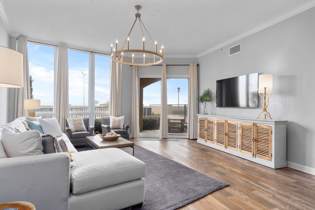 A light-filled living area features large windows offering views of the outdoors. A neutral sofa is positioned on a stylish area rug, complemented by a coffee table and decorative elements. Wooden cabinets and a modern television add visual interest to the space.
