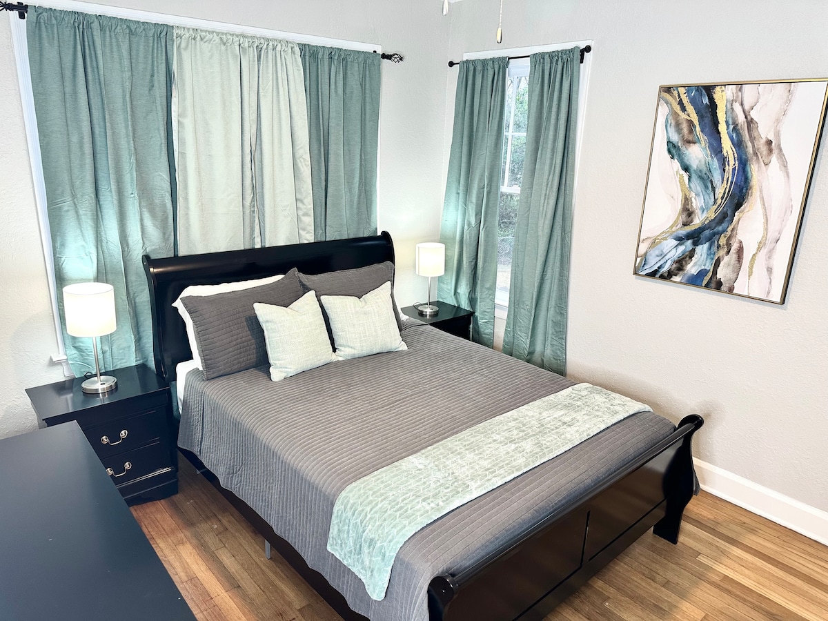 A welcoming bedroom features a dark wooden sleigh bed adorned with soft, neutral bedding and decorative pillows. Flanking the bed are two bedside tables with lamps. Light green curtains frame the windows, allowing natural light to brighten the room, while a modern artwork piece adds a touch of elegance.