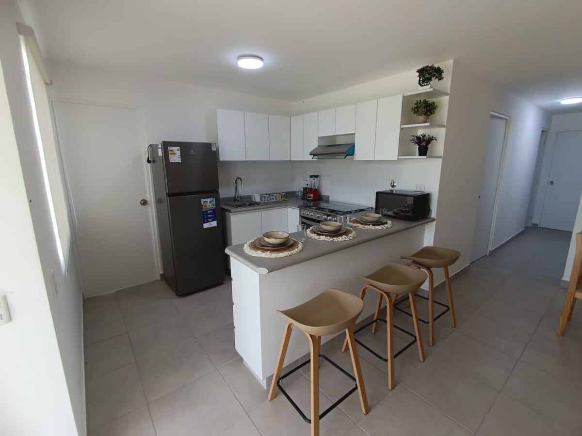 A modern kitchenette is presented, featuring a refrigerator, stove, and essential appliances. A central island with three minimalist stools offers seating. Neutral-toned cabinetry and light-colored walls enhance the open space, while natural light floods in from a nearby window.