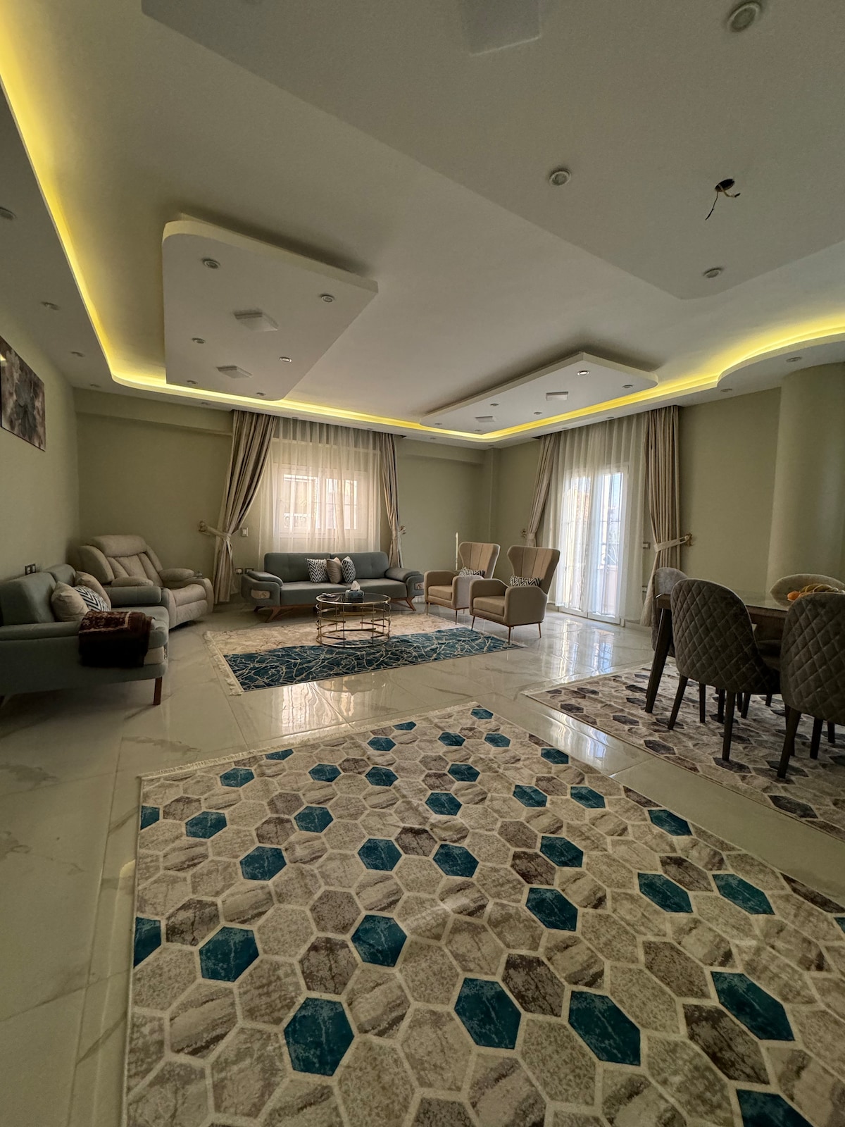 The living area features a blend of plush seating and elegant dining options. Soft lighting complements the neutral-toned walls. Decorative rugs in geometric patterns enhance the space, while large windows allow natural light to fill the room, creating an inviting atmosphere.