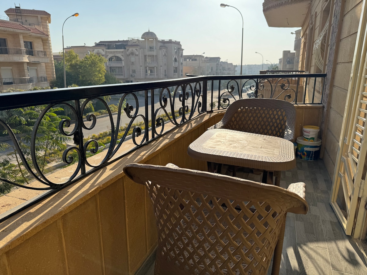 A balcony is presented with a wrought iron rail and a view of the surrounding neighborhood. Two brown wicker chairs are positioned around a small table, creating a space for relaxation. Sunlight illuminates the area, enhancing the visibility of the outdoor setting.