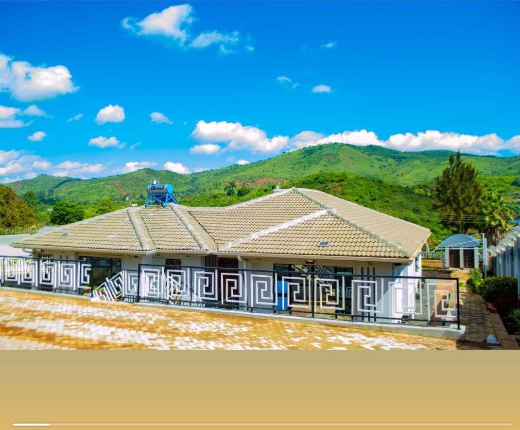 A single-story building is set against a backdrop of rolling green hills, featuring a distinctive roof and decorative exterior. Sunlight illuminates the structure, highlighting the architectural details, while a clear blue sky with scattered clouds enhances the overall ambiance.