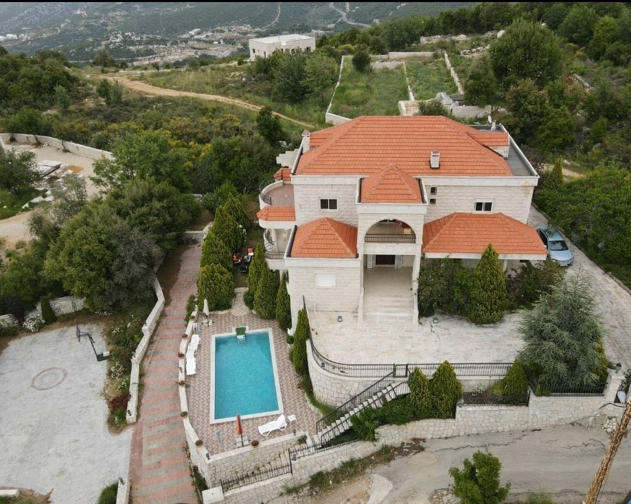 An aerial view showcases a spacious stone villa surrounded by lush greenery. A swimming pool is visible in the backyard, accompanied by sun loungers. The red-tiled roof and manicured landscaping create a serene atmosphere, while the nearby hills enhance the tranquil setting.