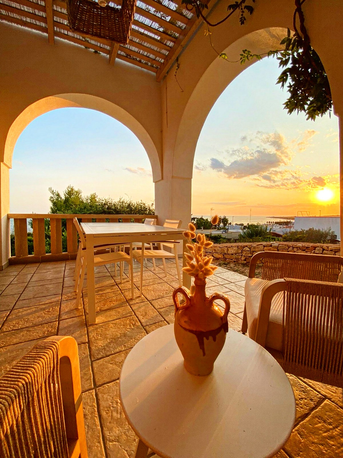 An outdoor seating area is highlighted by a table and chairs, framed by large arched openings that provide views of the sunset. A decorative vase sits on a small table, complemented by natural light filtering through trellised overhead beams.