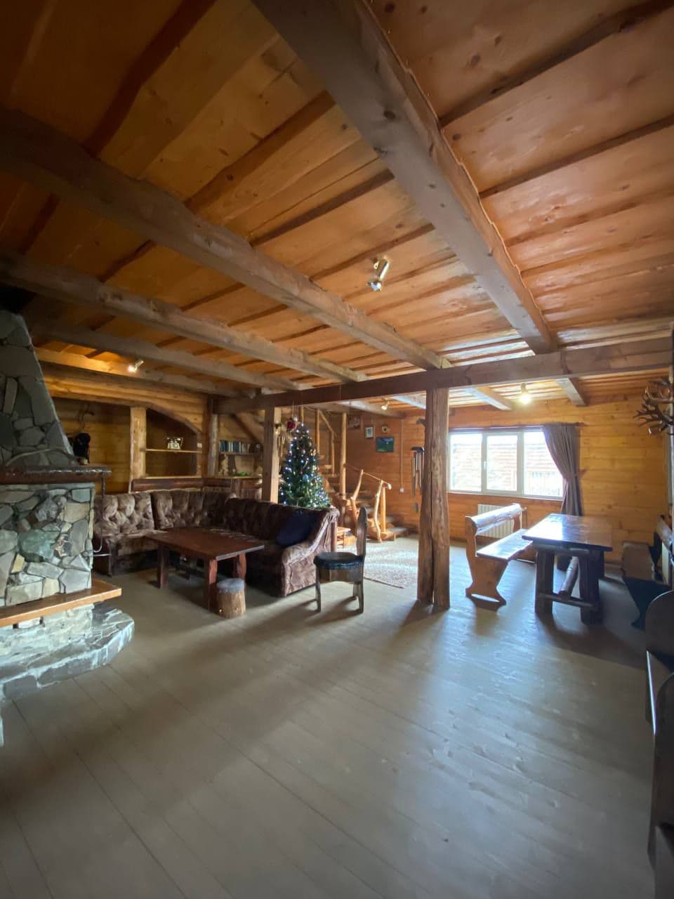 An interior space features warm wooden beams and a stone fireplace. A seating area is arranged with a brown tufted sofa and a large wooden table. Decorative elements include a holiday tree, while windows allow natural light to fill the room, illuminating the cozy atmosphere.