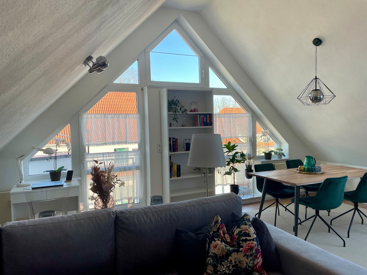 An airy open-concept living space features a large window allowing natural light to fill the room. A cozy sofa and a dining area with a wooden table and green chairs create a welcoming ambiance. Shelves with books and decorative items are visible, enhancing the room's character.