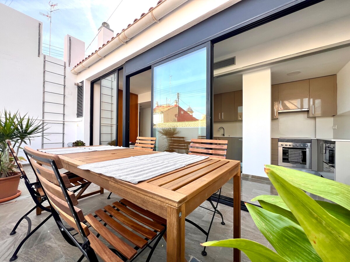 An outdoor dining area is shown, featuring a wooden table surrounded by several chairs. Large glass doors lead into a modern kitchen, and potted plants add a touch of greenery. Light reflects off the stone floor, enhancing the inviting space.