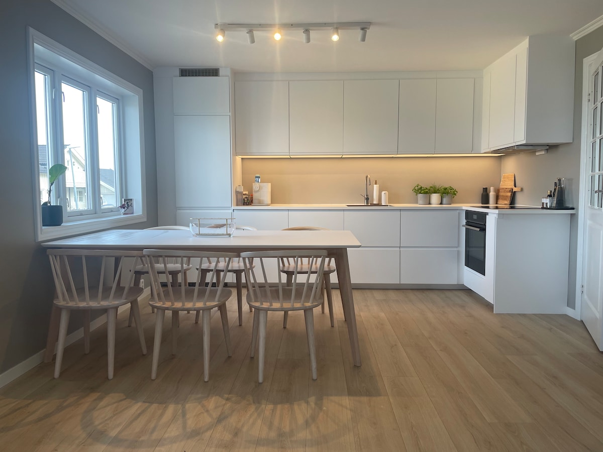 A modern kitchen features a spacious table surrounded by six light-colored chairs. Sleek cabinetry lines the walls, and warm LED lighting highlights the countertop. A window provides natural light, and potted plants add a touch of greenery to the space.