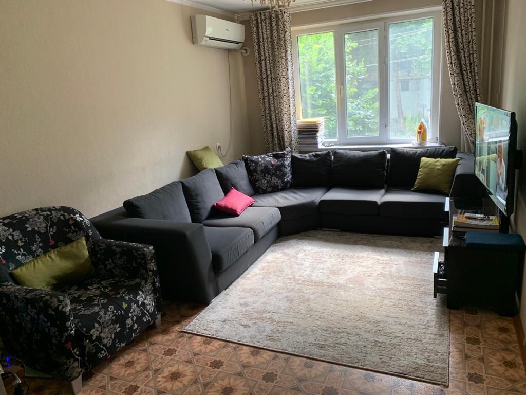 A spacious living area is furnished with a large sectional sofa in a dark color, accompanied by a floral-patterned chair. Colorful cushions add subtle accents. Natural light filters through a large window, illuminating the light-toned walls and the soft area rug underfoot.