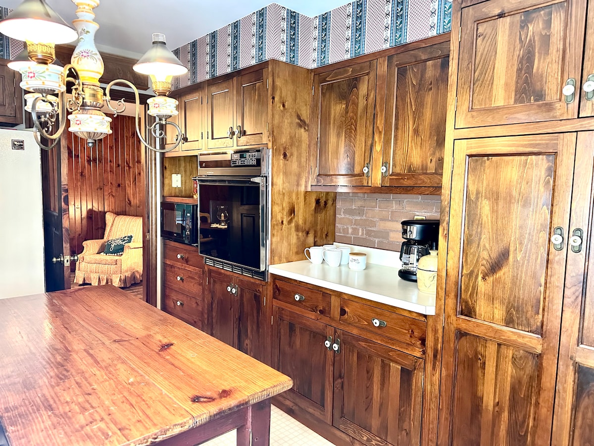 A warm-toned kitchen features wooden cabinetry and a central island. Modern appliances, including an oven and coffee maker, are integrated into the design. A vintage chandelier hangs above, and a cozy seating area with a chair is visible in the background.