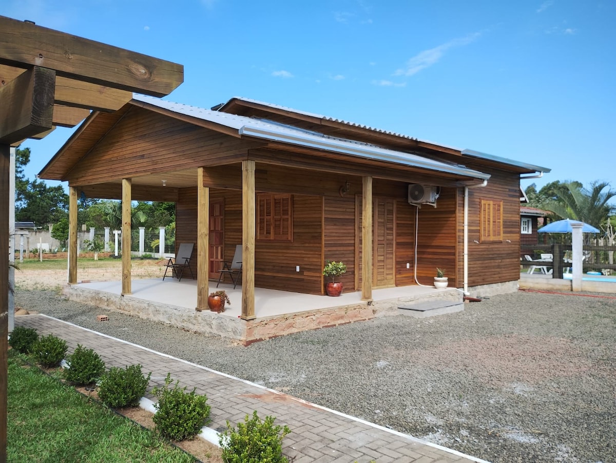 A wooden exterior is presented in a single-story structure, featuring a covered porch supported by wooden posts. Steps lead to the entrance, where two chairs are positioned. The surrounding yard is landscaped with small bushes and gravel pathways, with a pool visible in the distance.
