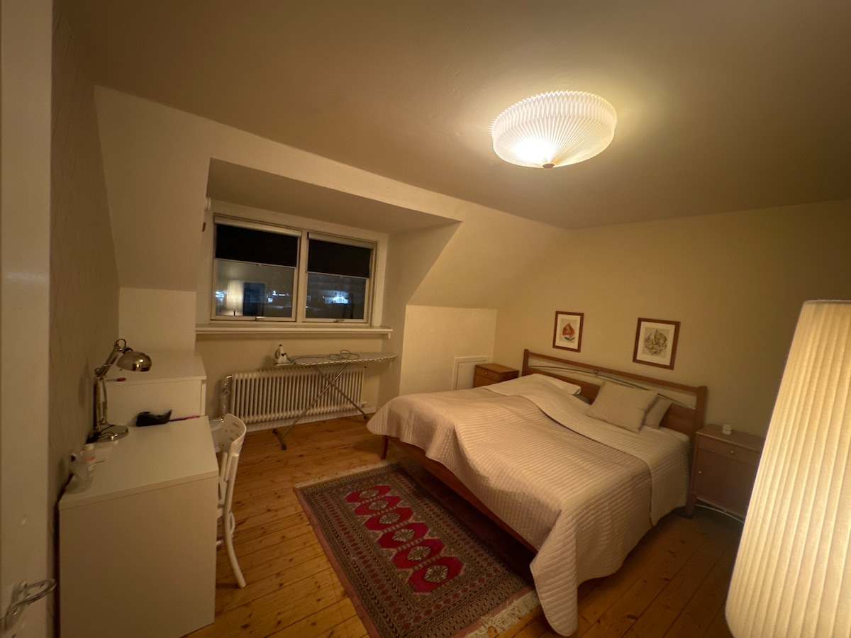 A bright bedroom features a spacious bed with light linens, framed artwork on the walls, and a wooden dresser. A window allows natural light to enter, and a desk with a chair is positioned nearby. A warm glow emanates from a standing lamp.