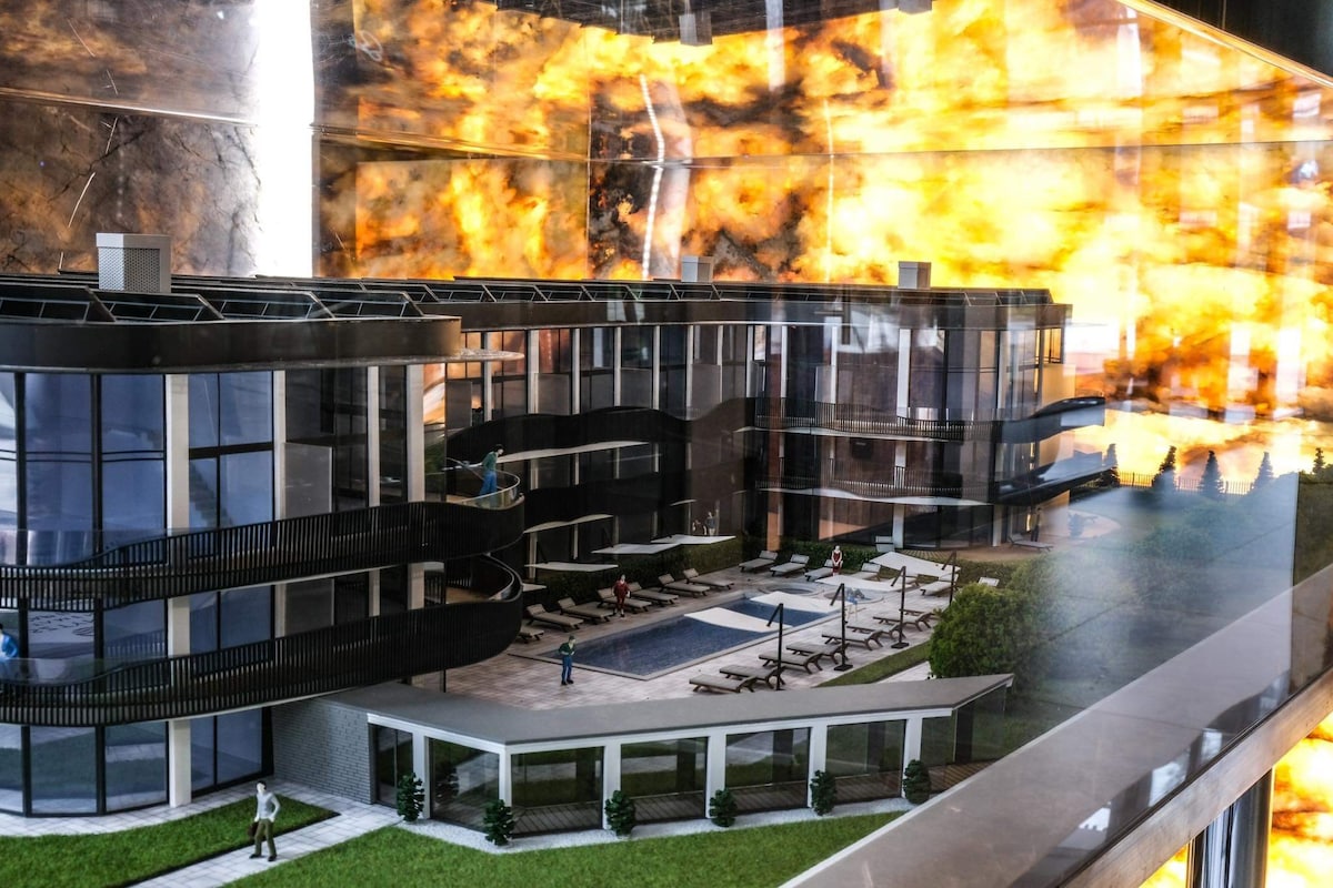 A detailed model of a modern apartment complex is presented, featuring multiple floors, large balconies, and a central outdoor pool area. Sun loungers surround the pool, framed by landscaped grass. Reflections and lighting enhance the visual depth of the structure within its display setting.