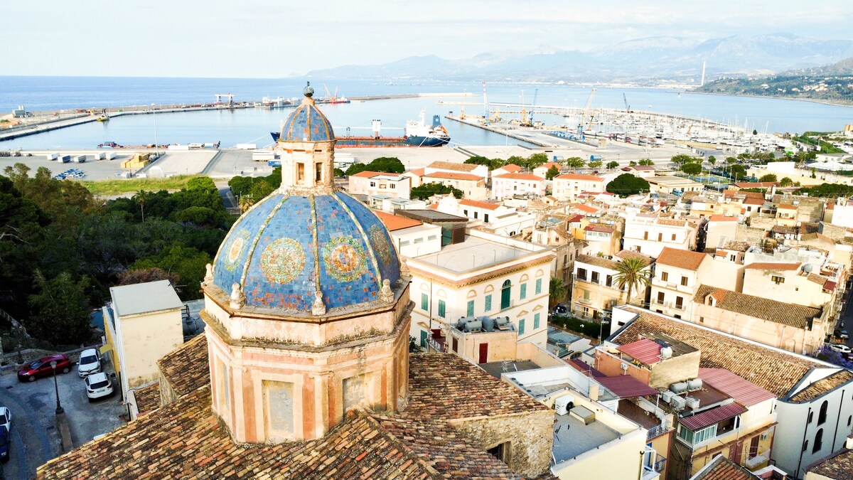 An aerial view highlights the vibrant dome of a historical building, adorned with intricate tile work. The background features the coastline, showcasing the marina and harbor filled with boats. The serene blue sea contrasts with the earthy tones of the surrounding buildings.