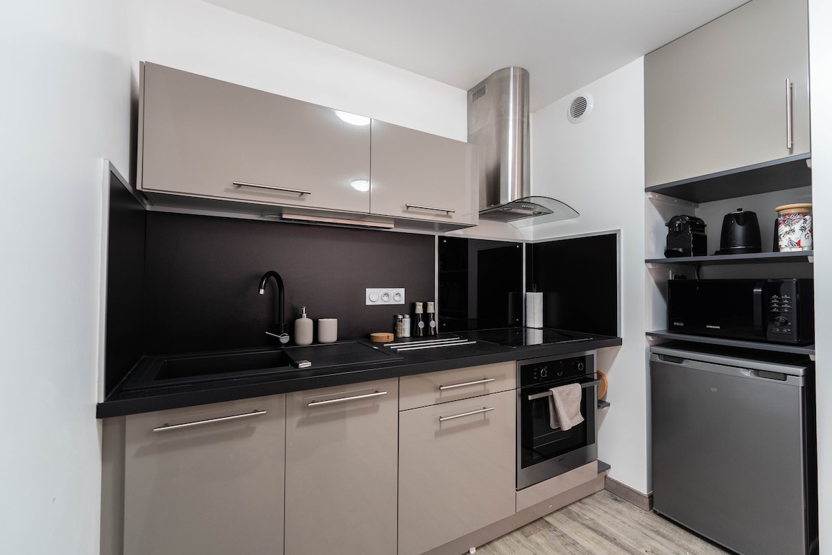 A modern kitchen features sleek cabinetry in neutral tones. The countertops are dark, complemented by a black backsplash. Appliances include a built-in oven, stovetop, microwave, and a compact refrigerator. Kitchenware and utensils are neatly organized on the counter.