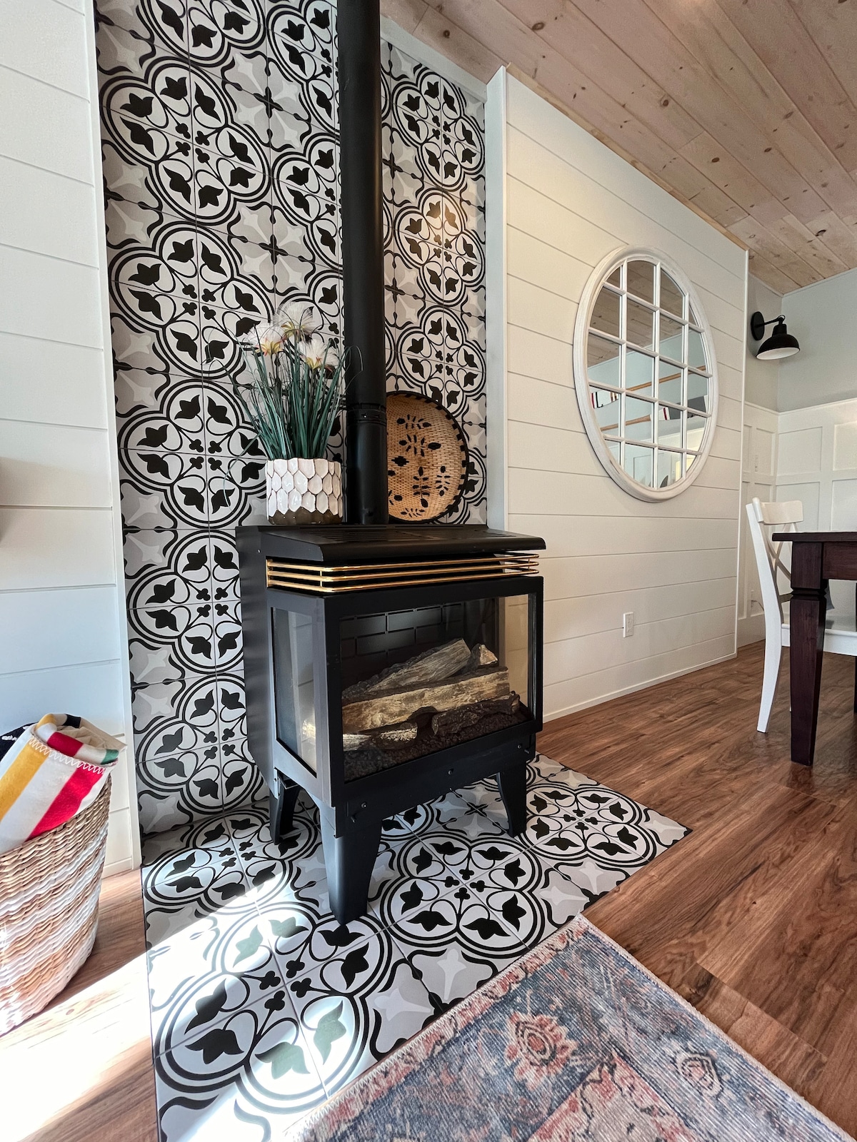 A stylish gas fireplace is positioned against a patterned feature wall, featuring intricate black and white design. A round mirror reflects light, while decorative plants sit atop the fireplace. A woven basket and an area rug add warmth to the space.