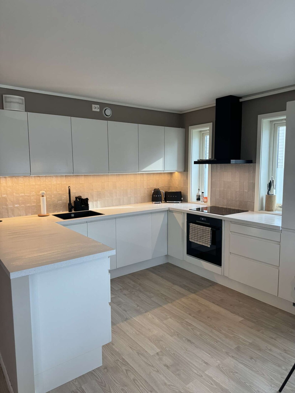 A modern kitchen is displayed with sleek white cabinetry and a minimalist design. Ample counter space is available, complemented by light-colored flooring. Large windows allow natural light to brighten the area, enhancing the functionality of appliances, including a built-in oven.