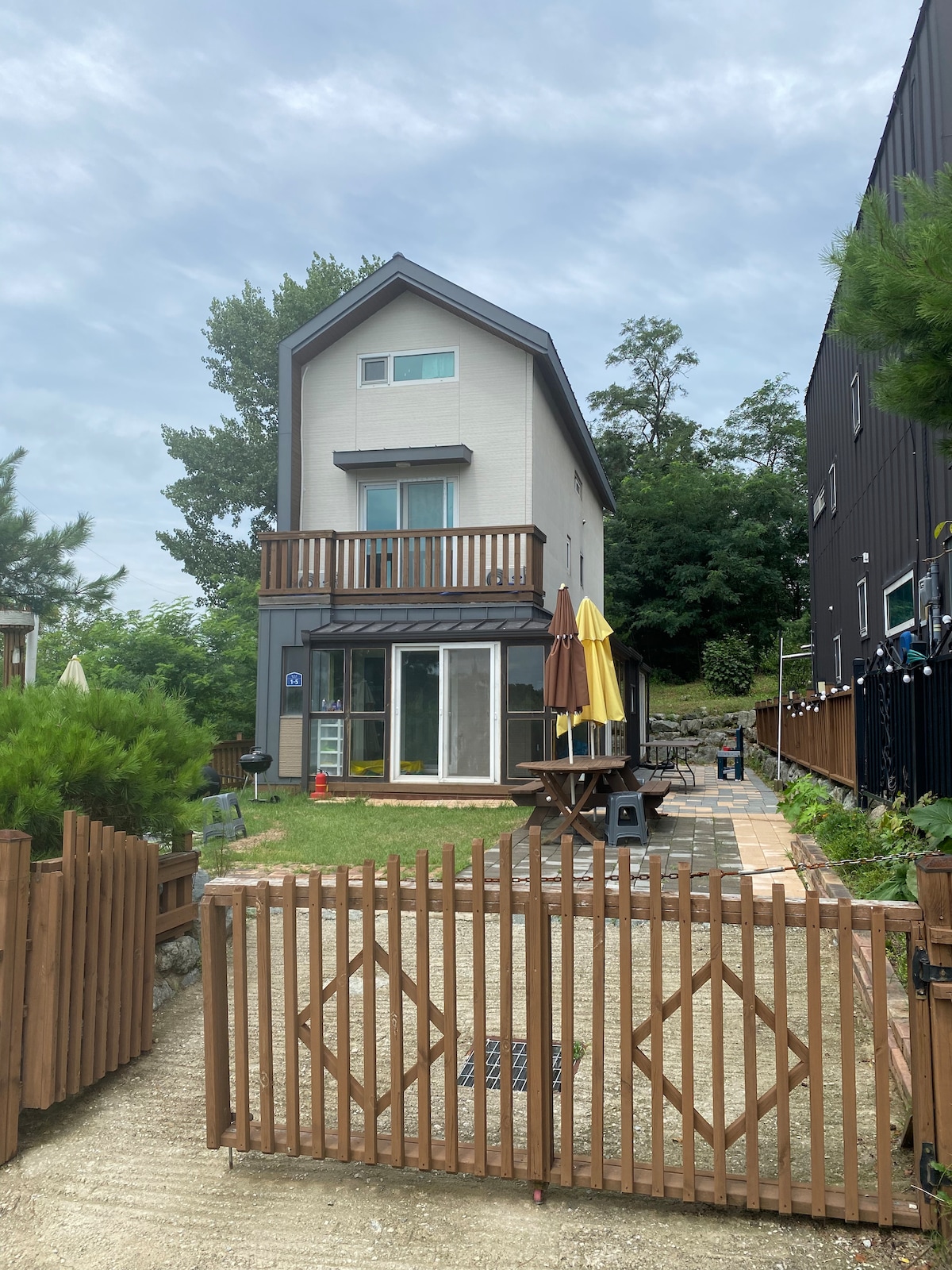 A three-story building is visible, featuring a light-colored exterior and a balcony on the second floor. The front yard is landscaped, with green grass, a patio area with umbrellas, and a wooden fence enclosing the space.
