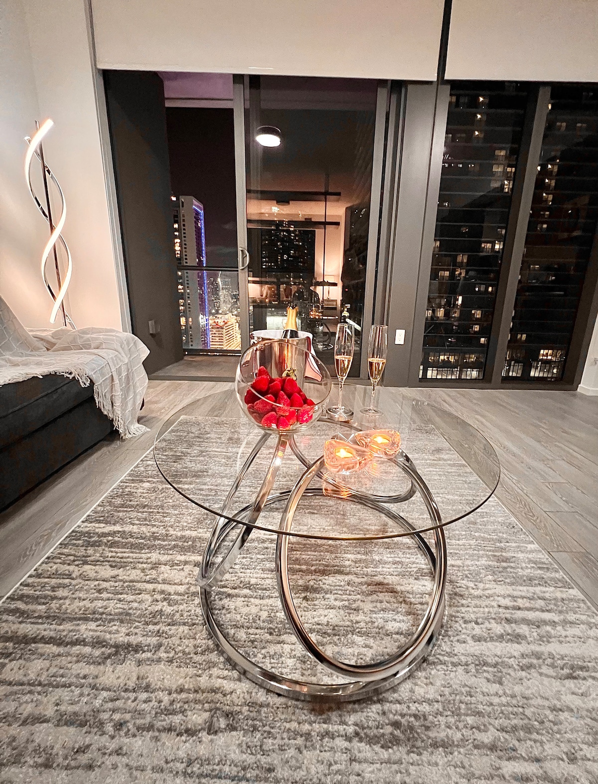 A modern living area features a sleek glass coffee table with a bowl of strawberries and champagne flutes, complemented by soft candlelight. Floor-to-ceiling windows provide views of the city skyline at dusk, with vibrant lights visible in the background.