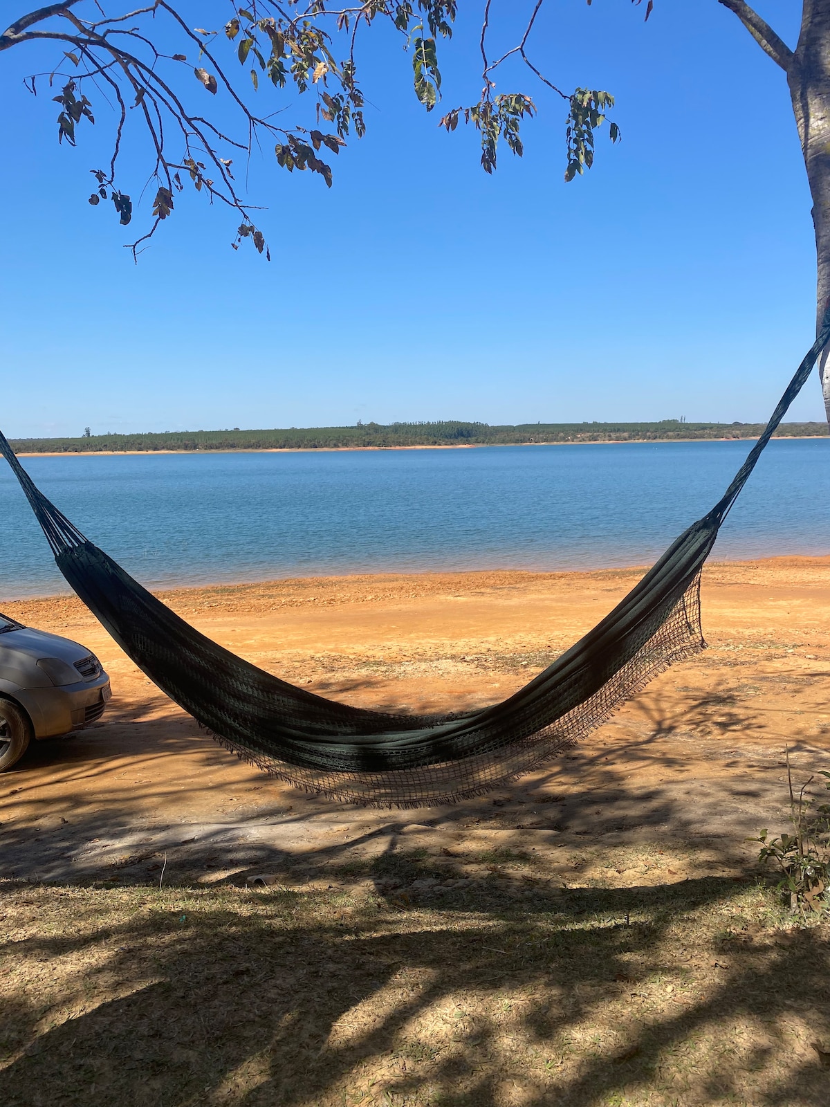 A hammock is suspended between two trees, overlooking a calm body of water. The shore features sandy earth tones, and distant greenery can be seen along the water's edge. A vehicle is parked nearby, suggesting accessibility to the tranquil spot.