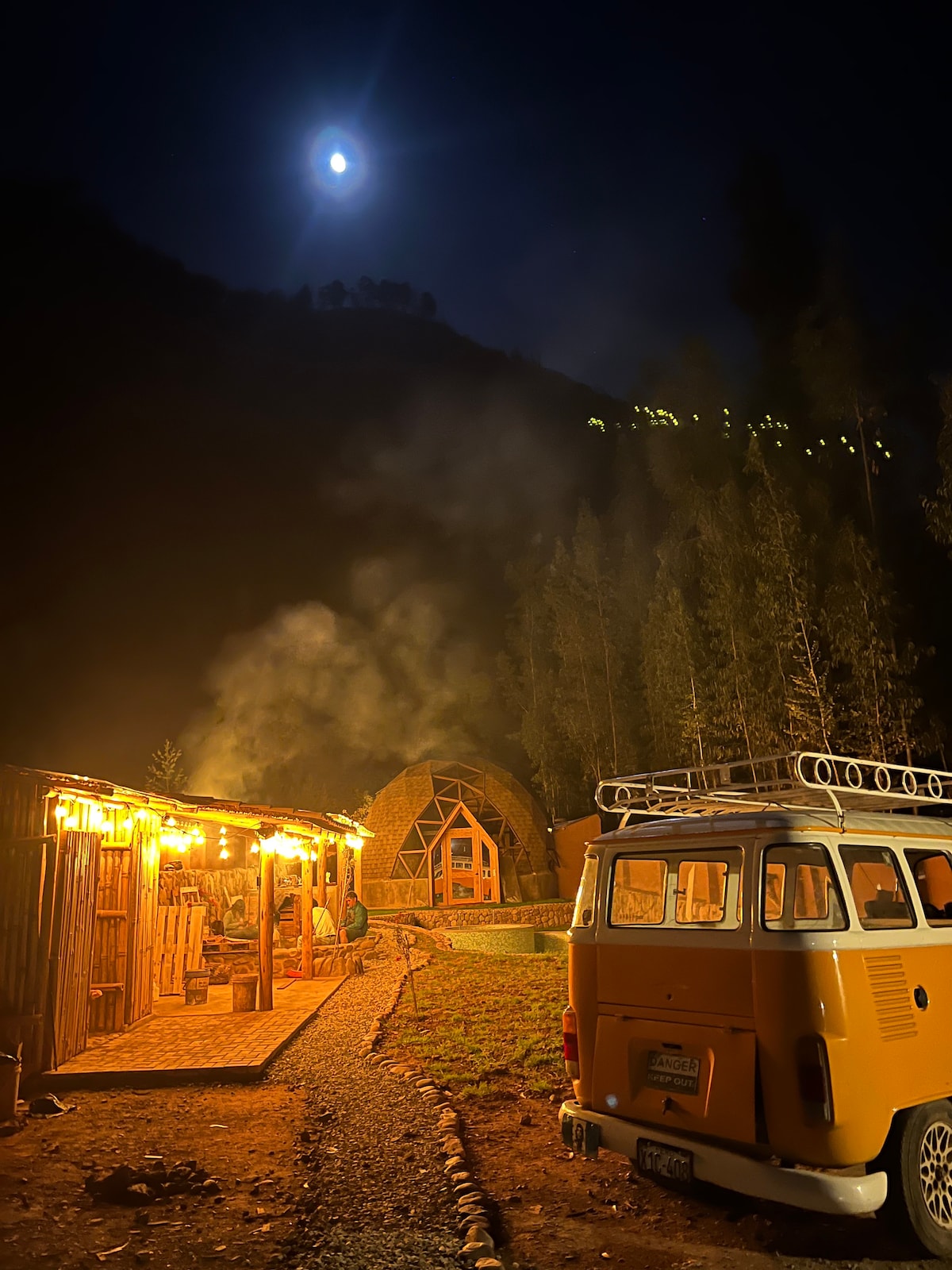 A vintage Volkswagen van is parked near a charming wooden structure, softly illuminated by string lights. In the background, a luxurious dome glows under the moonlight, surrounded by trees and hills. Smoke gently rises from the area, adding to the serene evening atmosphere.