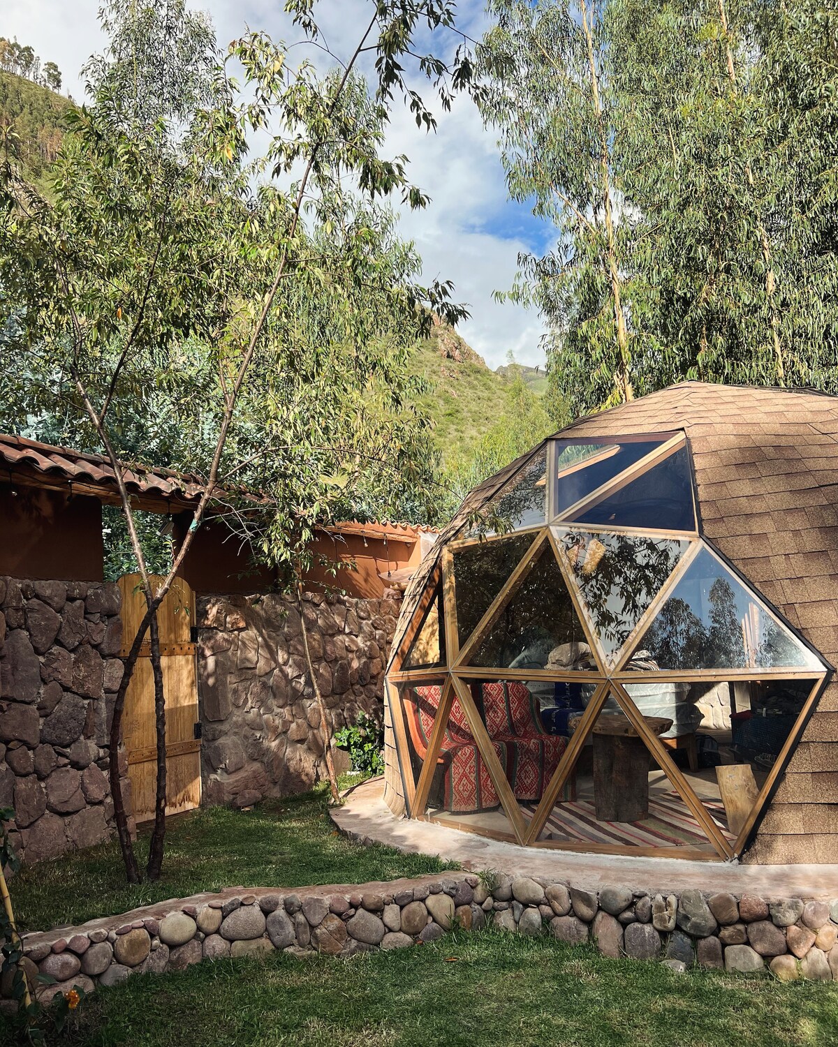 A modern geodesic dome features large glass panels surrounded by lush greenery. Natural stone walls enclose the area, enhancing the connection to nature. A grassy space provides a serene setting, complemented by trees that offer shade and privacy.