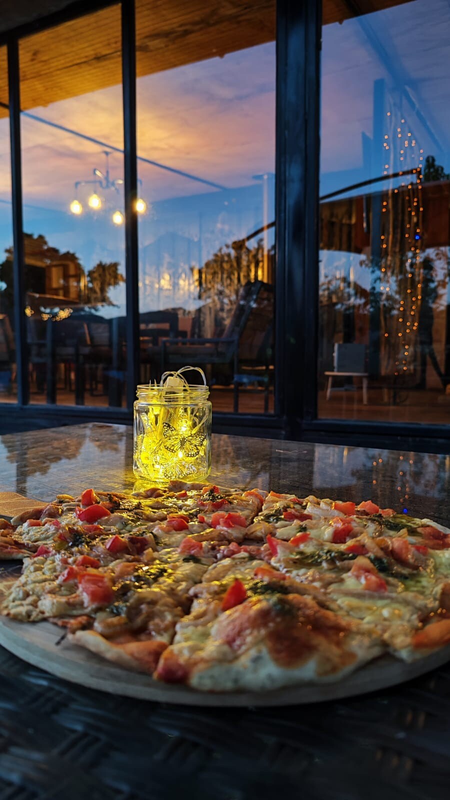 A wooden pizza board is displayed in the foreground, featuring a freshly made pizza topped with diced tomatoes and herbs. In the background, a softly lit dining area can be seen through large glass doors, with warm lights creating an inviting environment.
