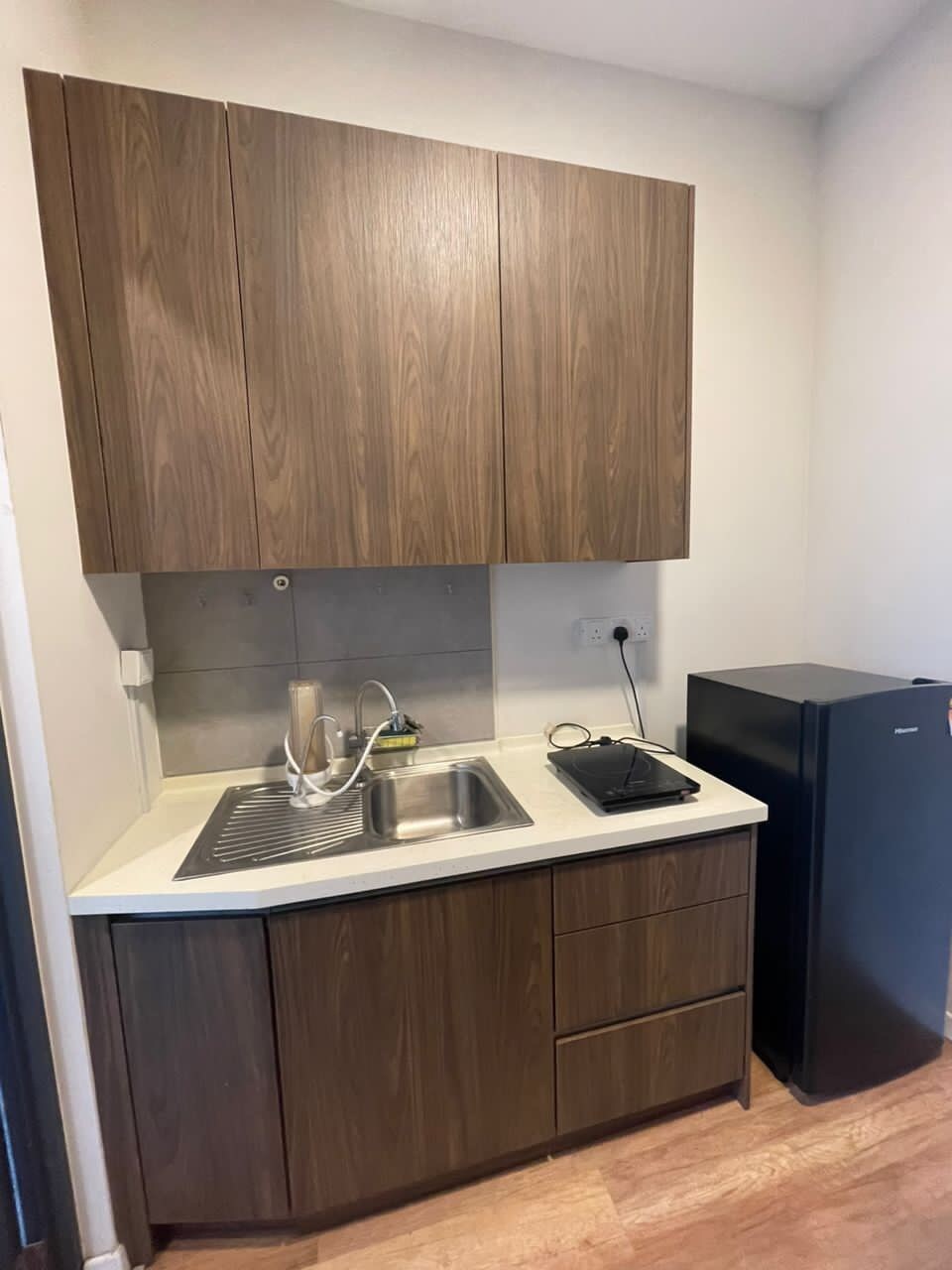 A compact kitchen area features wooden cabinetry and a white countertop. A sink is positioned beneath a wall-mounted cabinet. Nearby, a stovetop is visible alongside a small refrigerator, creating a functional space for meal preparation.