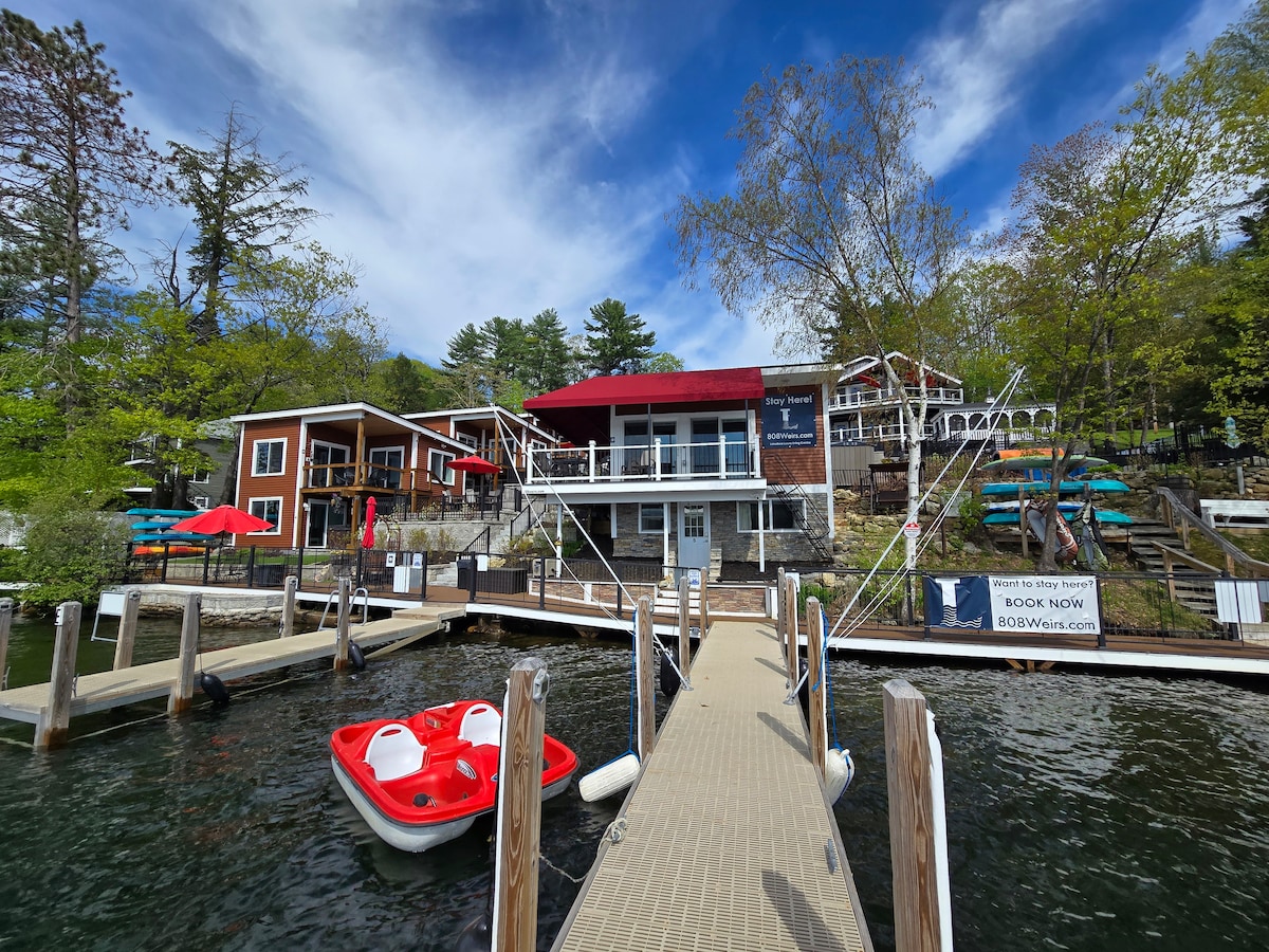 The waterfront cottage is observed from the dock, featuring a two-level structure with a red awning. A spacious deck is visible, with outdoor seating overlooking the lake. A small red paddle boat is docked at the water's edge, surrounded by lush greenery.