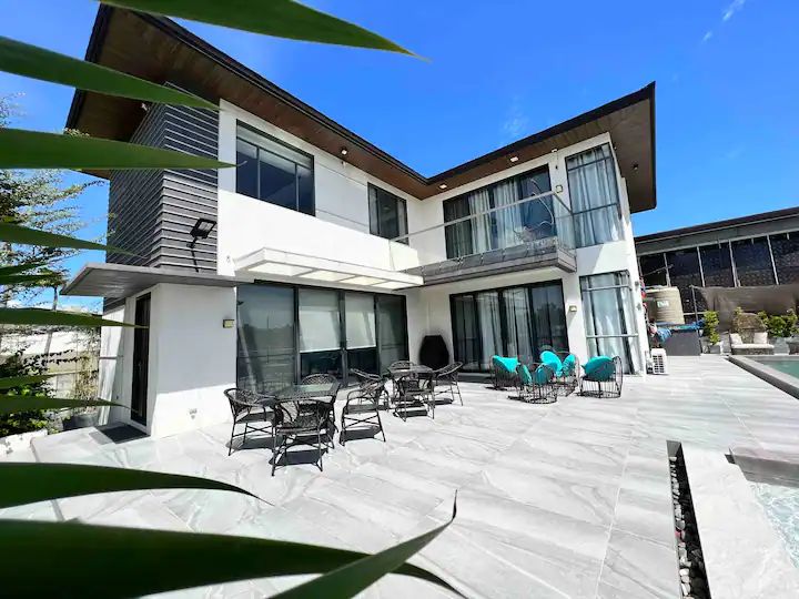 The exterior of the two-story home features a modern design, with large glass windows and a spacious outdoor patio furnished with several black wicker chairs and turquoise accent chairs. A private pool is visible beside the patio, set against a clear blue sky.