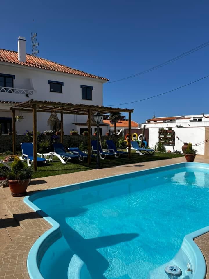 A bright outdoor space features a crystal-clear pool surrounded by lounge chairs and potted plants. A shaded area is created by a wooden pergola. The spacious house is visible in the background, set against a clear blue sky.