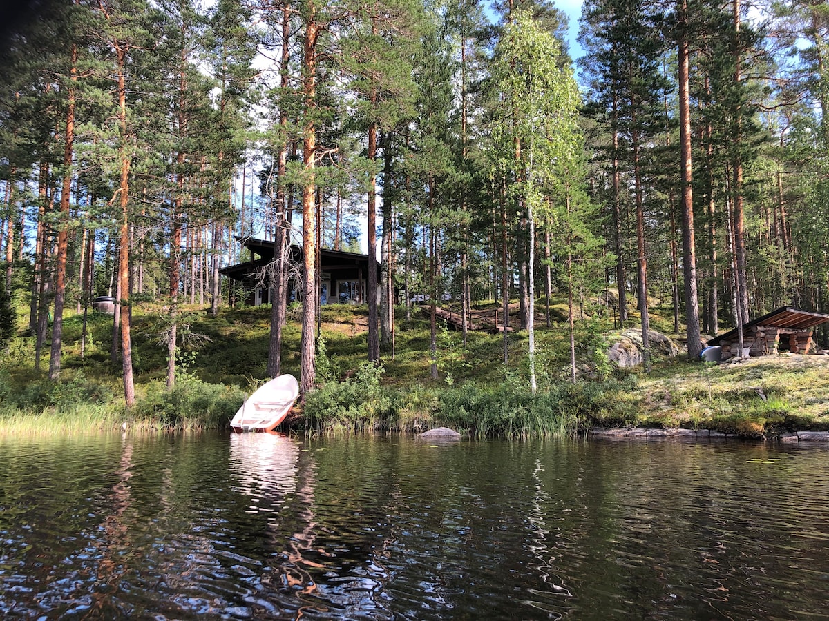 A modern cabin sits among tall trees on a gentle slope by the water's edge. The structure features large windows, allowing natural light to fill the interior. A small boat is anchored near the shore, surrounded by lush greenery and tranquil waters.