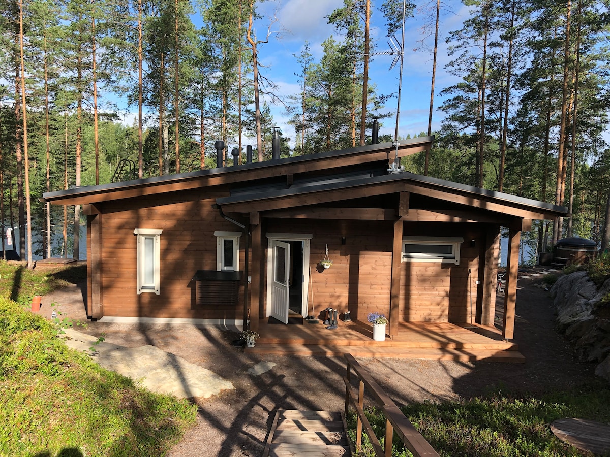 A modern wooden cabin with a sloping roof is nestled among tall trees. A welcoming front entrance is visible, featuring a small porch with steps leading up. Surrounding greenery highlights the cabin's natural setting, providing a serene view of the forest.