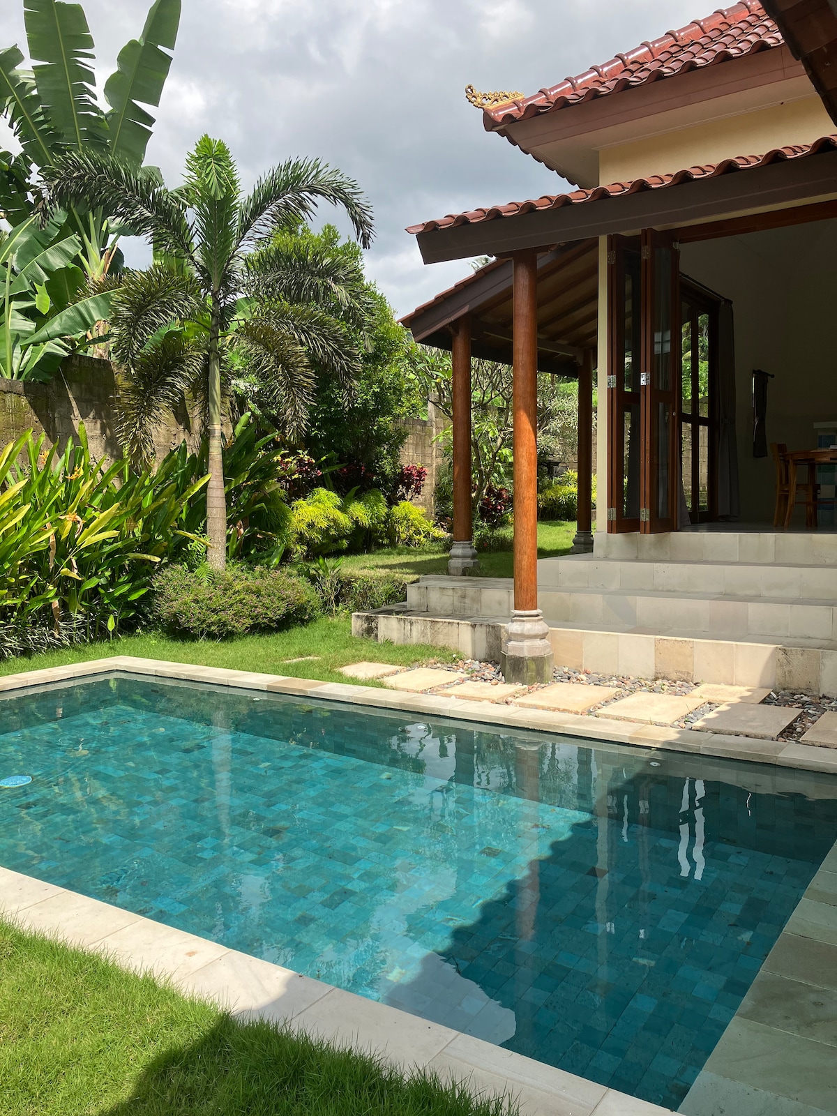 An inviting swimming pool is positioned in the foreground, featuring clear blue water surrounded by smooth stone tiles. Lush greenery, including palm and various tropical plants, frames the area. A traditional structure with wooden beams and inviting glass doors can be seen in the background.
