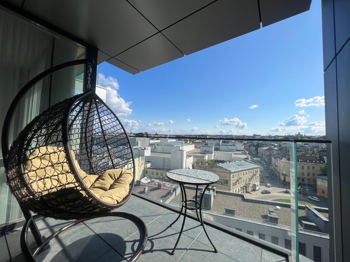 A hanging chair with a cushioned seat is positioned on a balcony, alongside a small round table. A cityscape is visible in the background under a clear blue sky, with clouds scattered overhead. The scene offers a view of surrounding rooftops and streets.