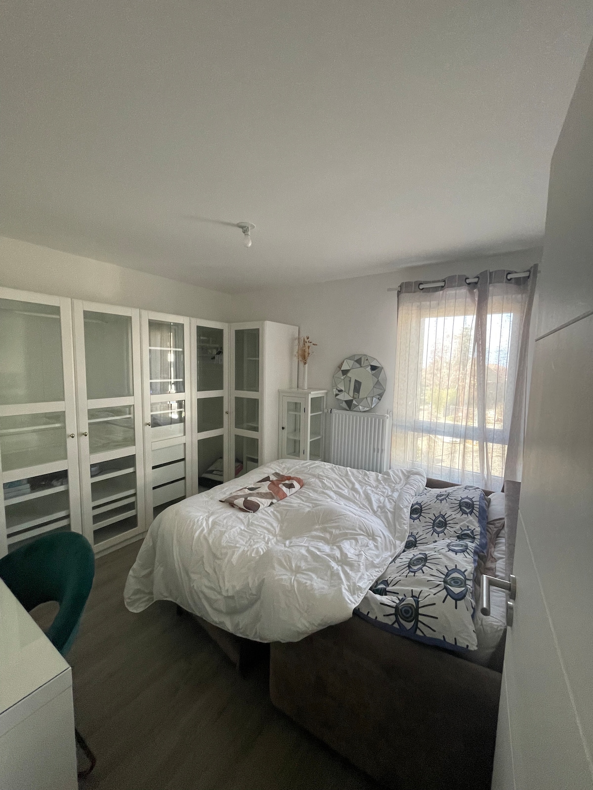 A bright bedroom showcases a spacious bed with white bedding, complemented by decorative cushions. Built-in wardrobes are situated along one wall, with a desk and chair nearby. Natural light filters through sheer curtains, creating a calming atmosphere in the room.