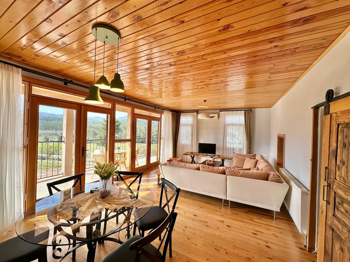 An open living area features a light wood ceiling and walls, enhancing a spacious feeling. A large sectional sofa is positioned near expansive windows, offering serene views of the surrounding nature. A round dining table occupies the adjacent space, set to accommodate guests comfortably.
