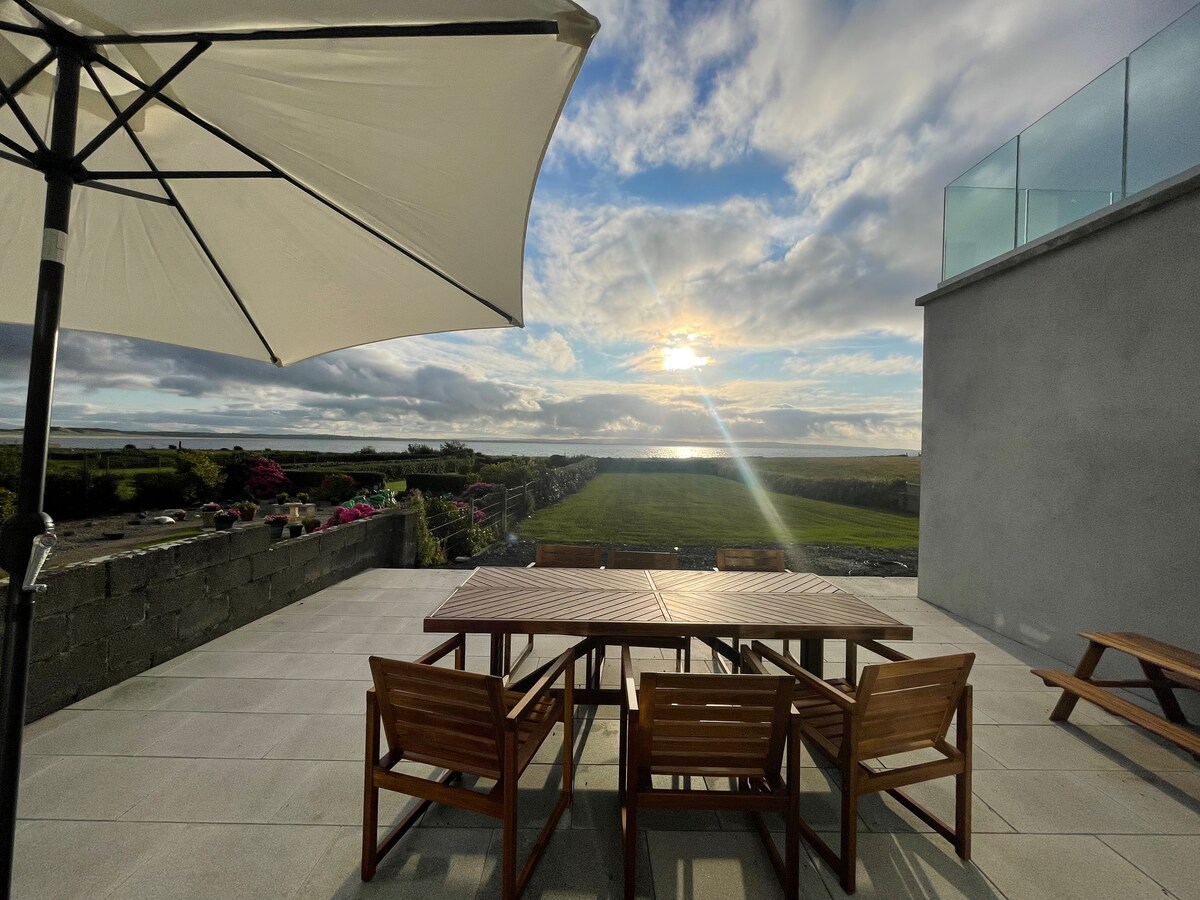 An outdoor terrace features a wooden dining table surrounded by six matching chairs, complemented by a large umbrella for shade. The scene captures a sweeping view of the coastline, with soft sunlight casting a warm glow over the landscape and clouds above.