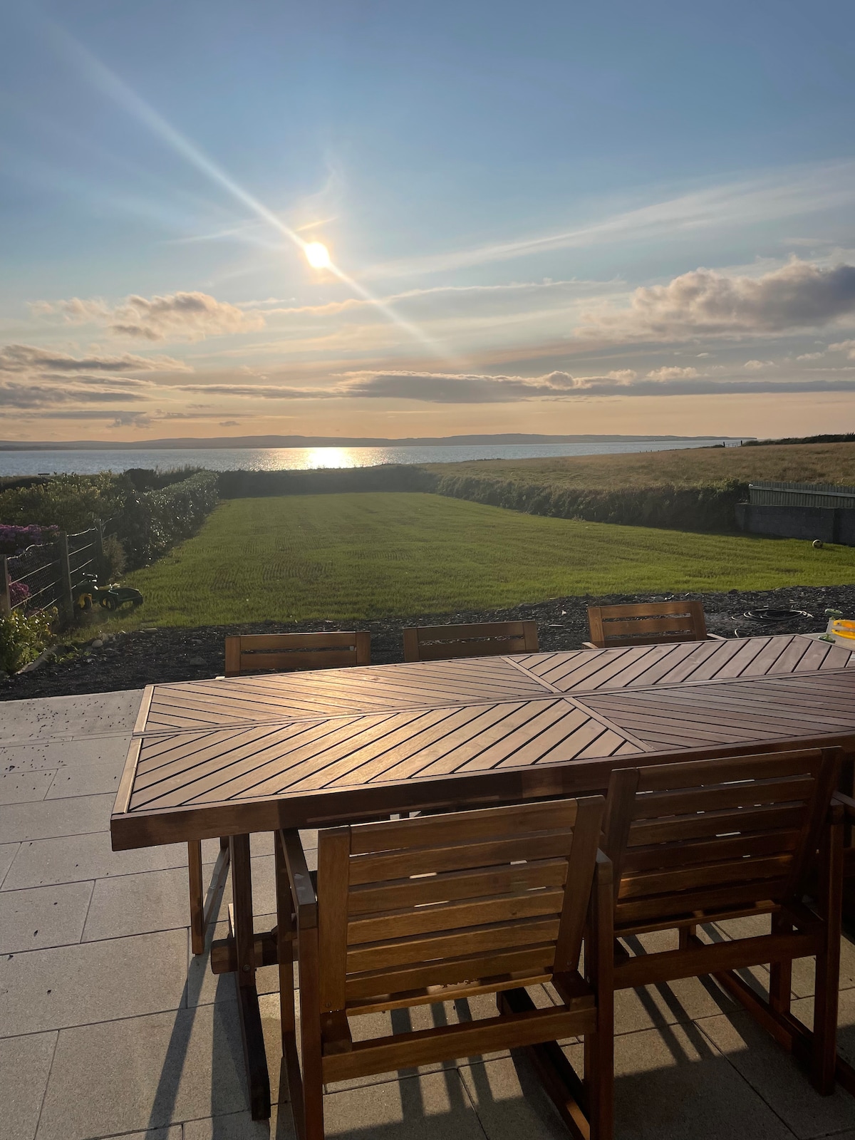 An outdoor dining area is shown, featuring a wooden table surrounded by matching chairs. The sun sets over a lush green lawn that extends toward the horizon, reflecting golden light on the peaceful water of the Atlantic Ocean.