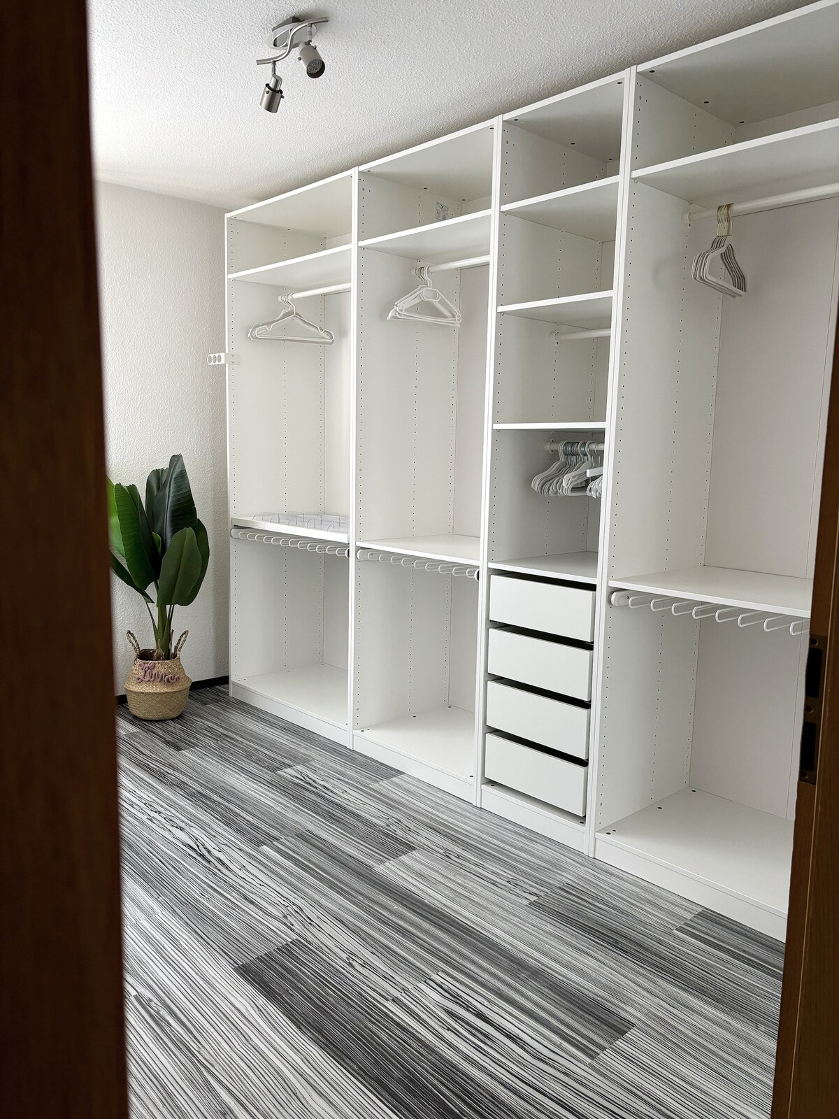 A well-organized walk-in closet features multiple shelves and hanging spaces. Light-colored cabinetry includes drawers for storage. A large potted plant adds a touch of greenery. The floor showcases a modern, striped design, contributing to the overall spacious feel of the area.