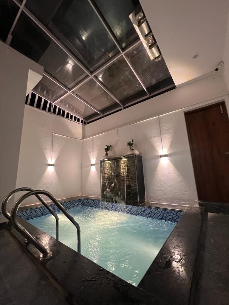 An indoor swimming pool is highlighted by a soothing water feature, with a waterfall cascading into clear water. Accent lighting on the walls creates a serene environment, while a glass ceiling allows natural light to flow into the space.